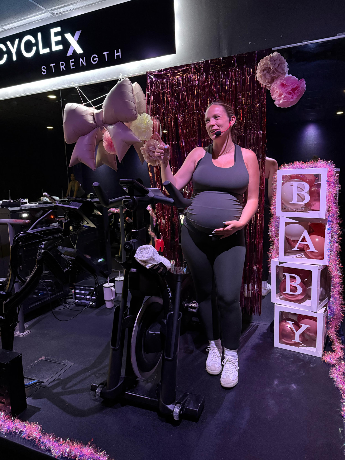 A smiling pregnant woman on the studio stage at a baby shower celebration, beside a decorated spin bike and stacked BABY letter blocks