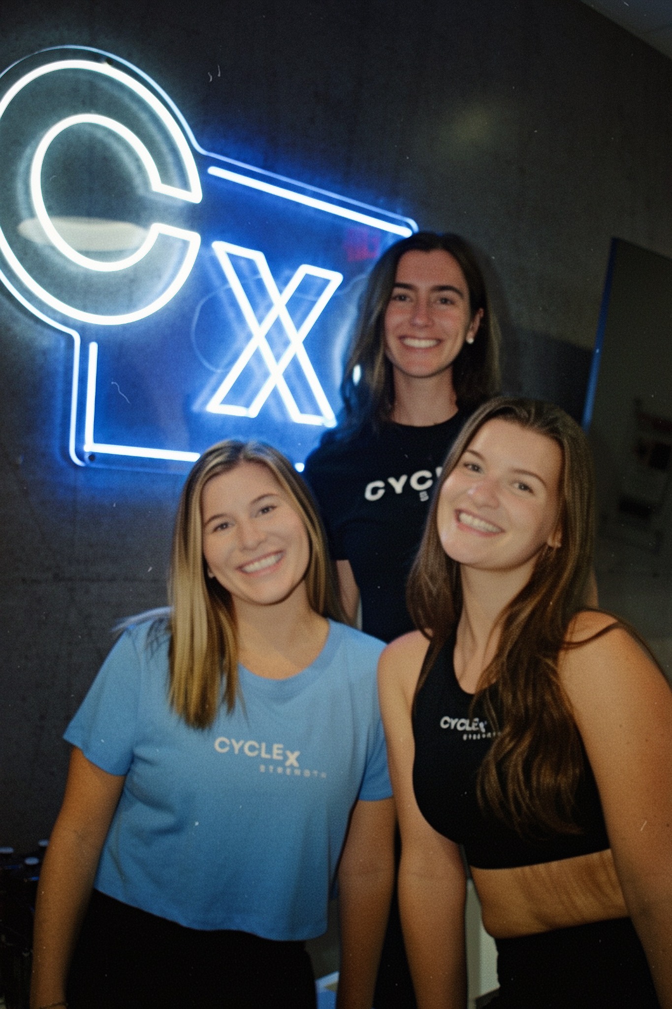 Three smiling members in Cycle x Strength gear in front of the studio's illuminated CxS sign