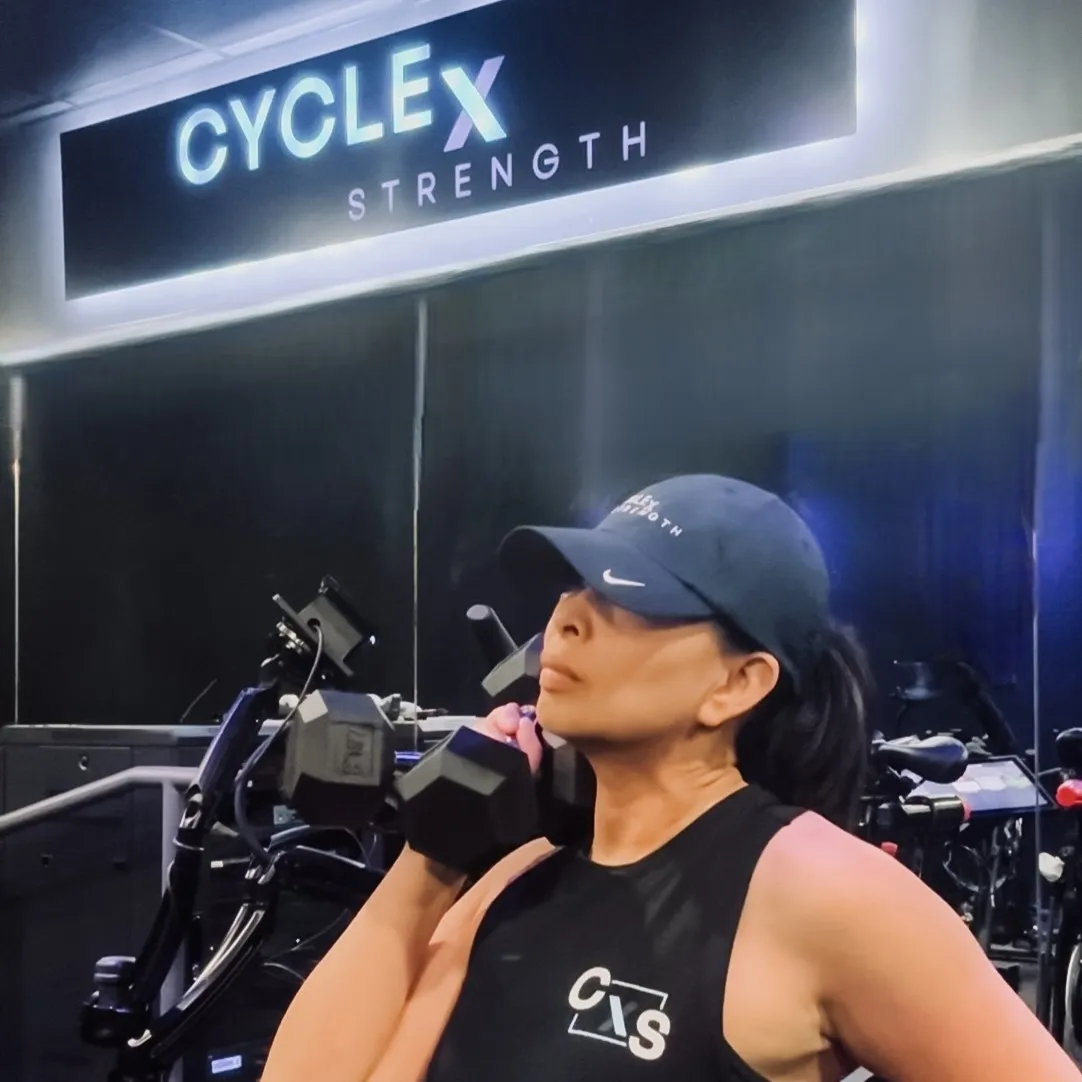 A member holding dumbbells beside the bikes under the Cycle x Strength sign