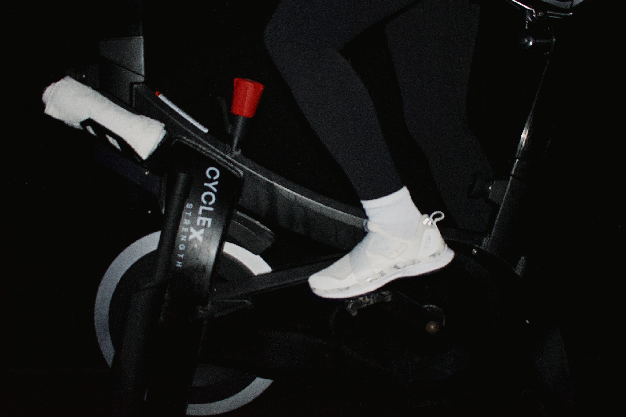 A white cycling shoe clipped into the pedal of a Cycle x Strength bike
